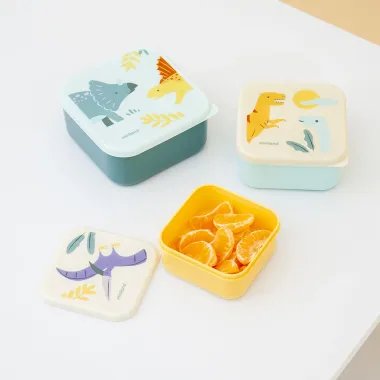 Miniland užkandžių dėžutės vaikams Dinozaurai sudėtos ant stalo, viena pripildyta apelsinų skiltelėmis