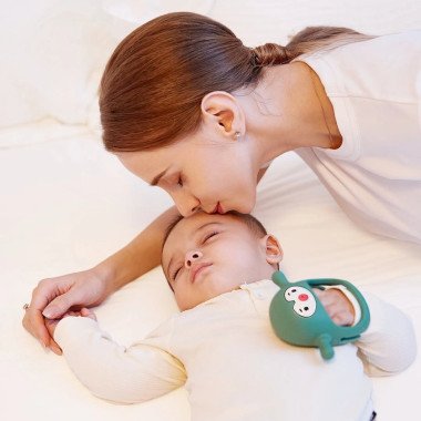 Mama bučiuoja miegantį kūdikį, kuris laiko Smily Mia silikoninį kramtuką